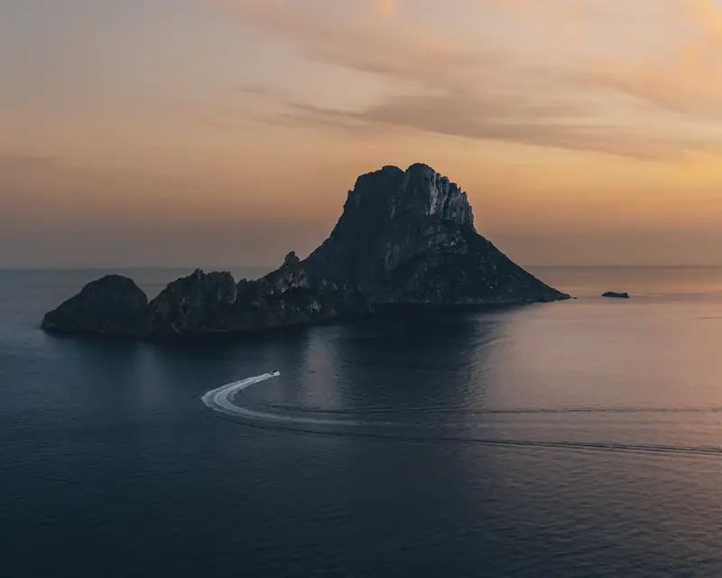 Una lancha deja su estela en el mar frente al islote de Es Vedrà, en Ibiza, al atardecer